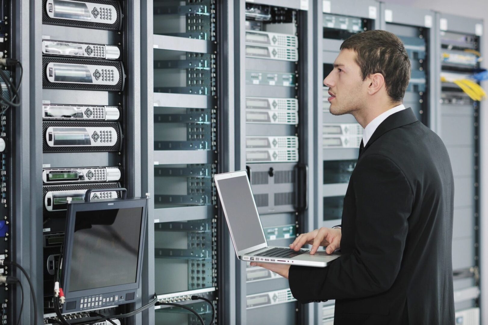 A man in black jacket holding laptop near wall of computers.